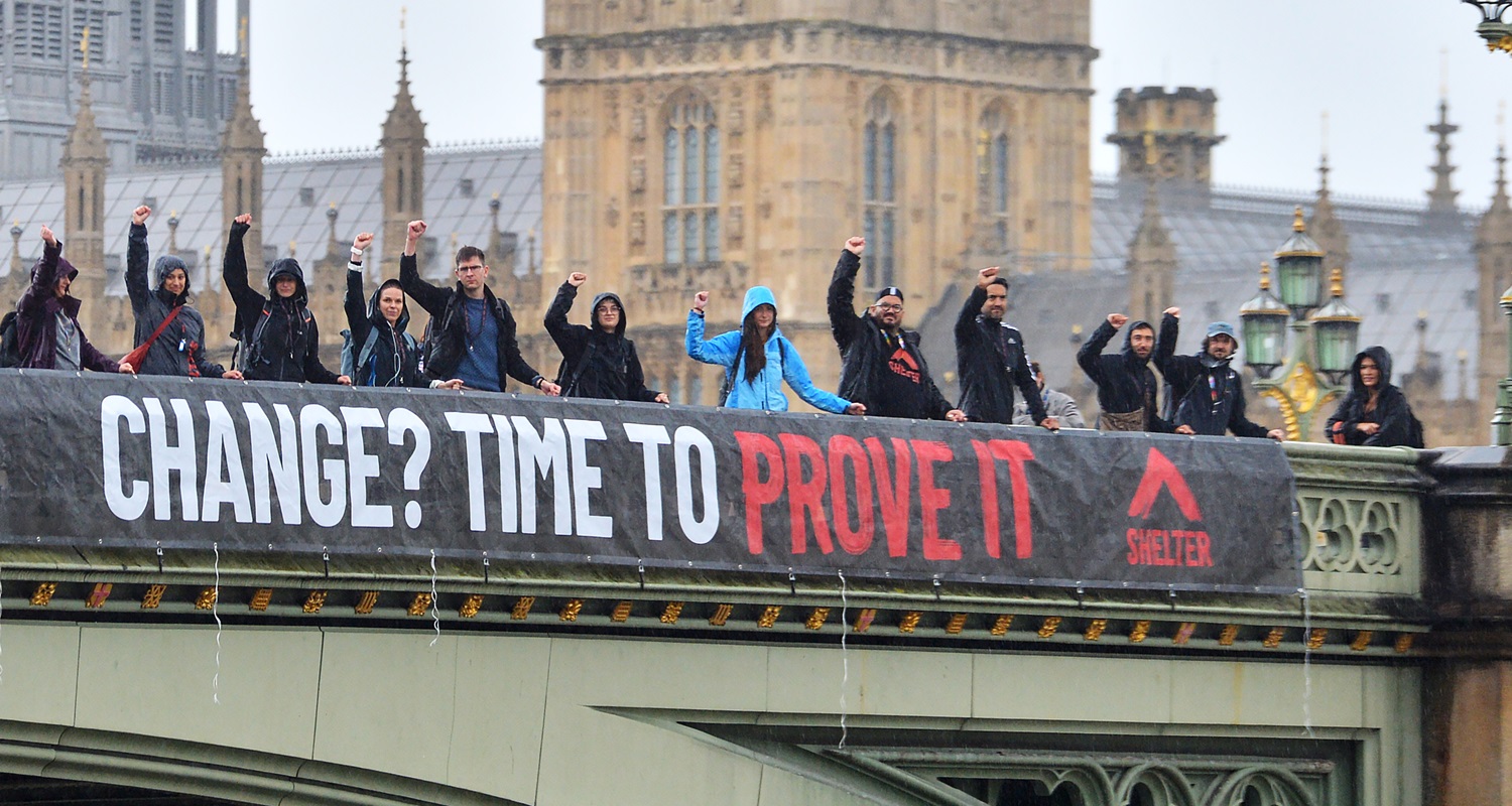 A group of campaigners have their arms held up high, and are stood hanging a banner from Westminster bridge which says "Change? Prove it!"