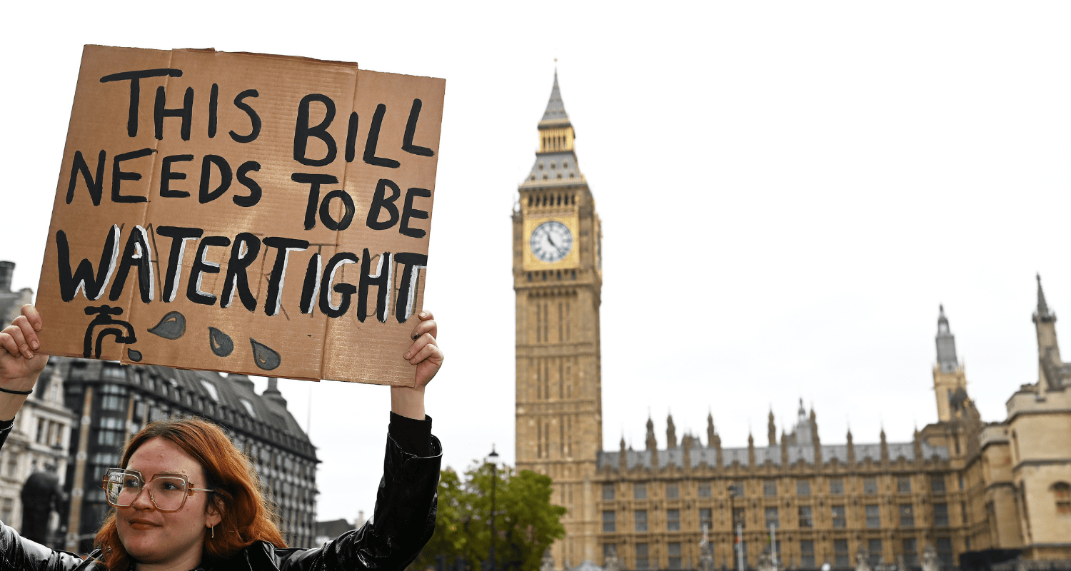 A woman stands holding a sign on the left hand side of the image. The sign reads 'This Bill needs to be watertight' with an image of a tap on it. Big Ben and parliament are in the background.