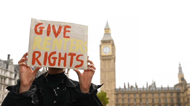 Someone holds a DIY placard over their face in Parliament Square. The sign reads 'Give Renters Rights' in orange and yellow paint.